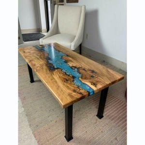 Puede incluir: Una mesa de centro rectangular de madera con un río de resina azul en el centro. La mesa tiene un acabado de vetas de madera natural y patas de metal negro. La mesa mide aproximadamente 122 cm de largo y 61 cm de ancho.