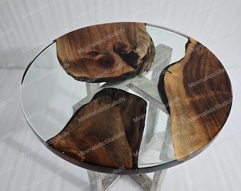 May include: A round side table featuring a clear glass top embedded with three distinct pieces of natural brown wood with visible grain patterns. The table is supported by a metallic base with a cross-shaped structure visible underneath the glass.