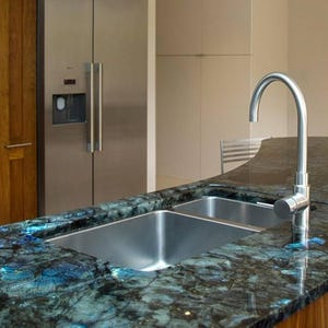 Könnte beinhalten: Moderne Kücheninsel mit einem Doppelwaschbecken aus Edelstahl und einem gebogenen Chromhahn. Die Arbeitsplatte ist dunkler Granit mit blauen und grünen Adern. Holzschränke und ein Kühlschrank aus Edelstahl sind im Hintergrund.