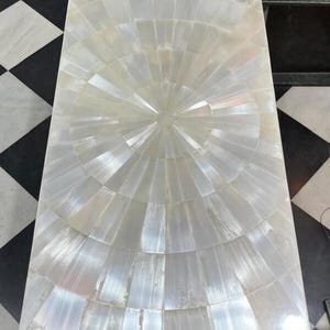 May include: Rectangular white coffee table with a mother-of-pearl inlaid surface. The table's surface features a radial design, with the iridescent shell pieces arranged in a sunburst pattern. The table is set against a black and white checkered floor.