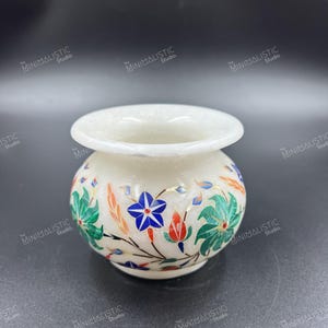 May include: A small, white marble vase with a rounded body and a wide opening. The vase is adorned with colorful floral designs, including blue, green, and red accents. The overall design is intricate and detailed, showcasing a blend of natural stone and artistic craftsmanship.