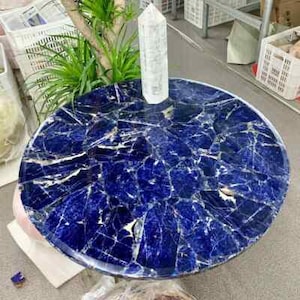 May include: A round table with a mosaic top in shades of deep blue and white. A clear crystal point stands upright on the table's surface. The table is surrounded by greenery and storage containers.