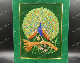 Puede incluir: Un bordado de pavo real enmarcado. El pavo real tiene un cuerpo azul, plumas de cola naranjas y azules, y está posado en una rama. El bordado está sobre un fondo de terciopelo verde con un borde dorado.