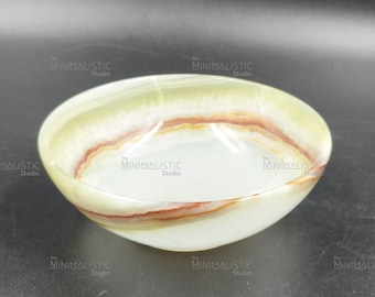 May include: A handcrafted, oval-shaped bowl with a translucent, polished finish. The bowl features layers of white, green, and reddish-brown hues, creating a natural, organic aesthetic. The Minimalistic Studio logo is visible.