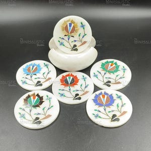 May include: Set of six white marble coasters with inlaid floral designs in various colors. Each coaster features a different flower design with blue, red, green, and orange petals. The coasters are round and come with a matching holder.