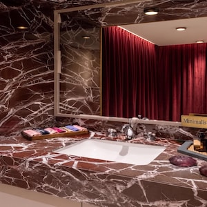 Puede incluir: Un tocador de baño con encimera y salpicadero de mármol rojo intenso con vetas blancas. Un lavabo blanco rectangular está centrado, acompañado por una bandeja de madera con paños plegados, un letrero dorado MinimalisticStudioIN y botellas oscuras.