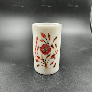 Könnte beinhalten: Ein weißer Marmorbecher mit floralem Design. Der Becher hat eine rote Blume und rote und grüne Blätter. Das Design ist in den Marmor eingelegt. Der Becher ist zylindrisch und hat eine glatte Oberfläche.
