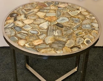 May include: Round side table with a tabletop made of polished agate stones in shades of gray, beige, and gold. The tabletop is framed by a silver metal edge and supported by silver metal legs.