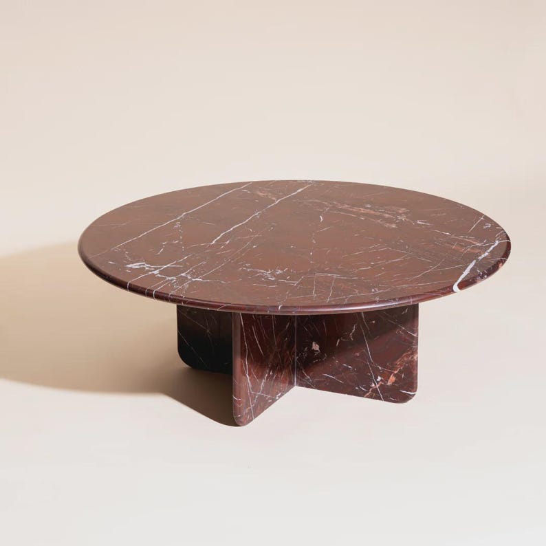 May include: A round coffee table with a deep red marble top and a cross-shaped base. The marble has white veining throughout. The table is a modern design and would be a statement piece in any living room.