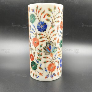 May include: A cylindrical white marble vase with intricate floral inlay. The design features colorful flowers in red, blue, green, and orange, with detailed leaf patterns. The vase has a smooth, polished finish and a minimalist aesthetic.