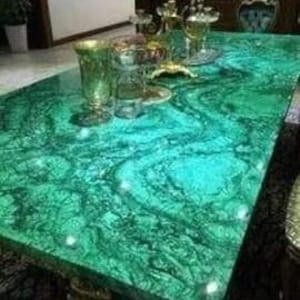 Può includere: Un tavolo rettangolare verde smeraldo con un motivo a vortice, possibilmente malachite. Il tavolo ha una superficie lucida ed è adornato con vetreria decorativa e un candelabro. Le dimensioni del tavolo non sono visibili.