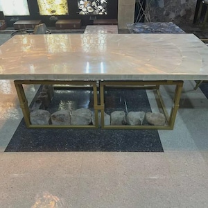 Puede incluir: Una mesa de centro rectangular con una tapa clara y estampada. La base de la mesa es un marco de metal dorado y presenta una fila de piedras en el interior. La mesa está colocada sobre un suelo con un patrón en blanco y negro.