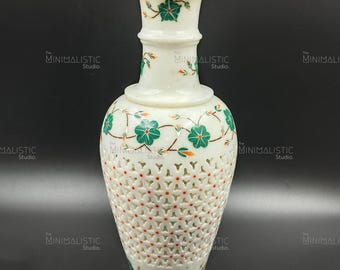 Könnte beinhalten: Eine weiße Marmorvase mit floralem Design. Die Vase hat einen breiten Sockel, einen gitterartigen Mittelteil und eine ausgestellte Oberseite. Grüne und orangefarbene florale Akzente verzieren die Vase. Die Vase ist etwa 30 cm hoch.