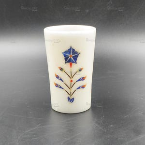 May include: A white marble tumbler with a floral inlay design. The design features a blue star-shaped flower with red accents and colorful leaves. The tumbler is cylindrical and stands on a dark surface.