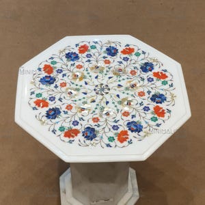 May include: White marble side table with an octagonal top. The tabletop features a floral inlay design with red, blue, and green accents. The base is a geometric shape. The text "MINIMALISTIC Studio" is visible.