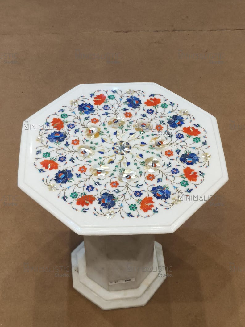 May include: White marble side table with an octagonal top. The tabletop features a floral inlay design with red, blue, and green accents. The base is a geometric shape. The text "MINIMALISTIC Studio" is visible.