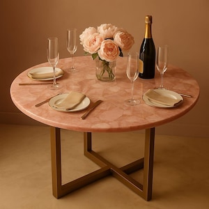 Pode incluir: Uma mesa redonda com um tampo de pedra de quartzo rosa polido e uma base de metal dourado. A mesa está posta com quatro taças de vidro transparente, dois pratos brancos com guardanapos bege dobrados, talheres dourados e um vaso com flores.
