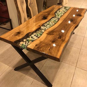 May include: A handcrafted wooden table with a river-like design. The table features a clear epoxy resin center filled with small stones and greenery. The wooden surface has a natural grain and is supported by black metal legs.