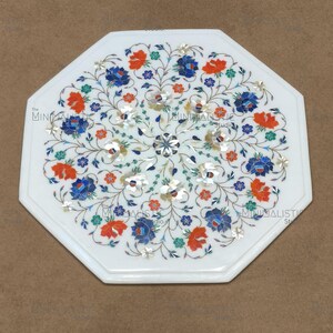 May include: White octagonal marble table with inlaid floral design. The intricate pattern features blue, orange, and silver-coloured flowers and vines. The table has a polished surface and a decorative edge.