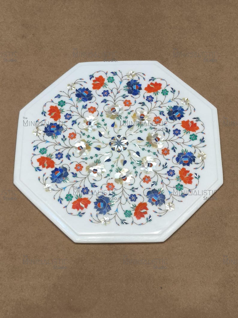 May include: White octagonal marble table with inlaid floral design. The intricate pattern features blue, orange, and silver-coloured flowers and vines. The table has a polished surface and a decorative edge.