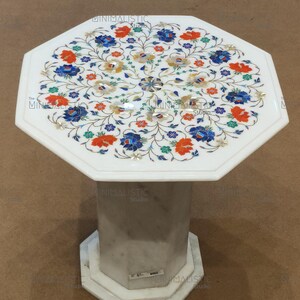 May include: White marble side table with an octagonal top and base. The tabletop features a floral inlay design with blue, orange, and yellow accents. The table has a single, octagonal pedestal base.