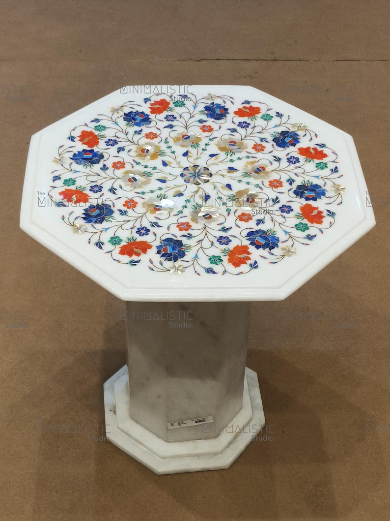 May include: White marble side table with an octagonal top and base. The tabletop features a floral inlay design with blue, orange, and yellow accents. The table has a single, octagonal pedestal base.