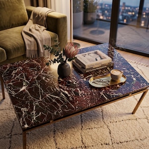 May include: A rectangular coffee table with a red marble top and gold-coloured metal legs. The table holds a tray with a coffee cup and glasses, books, and a vase with flowers. The table is in a living room setting.