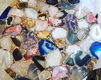 May include: A circular table top featuring a mosaic of polished, multi-colored agate slices in shades of blue, pink, white, gray, and black, embedded in a clear resin base with gold-toned metallic edges surrounding each individual stone piece.