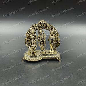 Op de afbeelding: Een goudkleurig religieus beeld met drie figuren in een boogvormig frame. De figuren staan op een decoratieve basis. Het beeld is gemaakt van metaal en heeft ingewikkelde details.