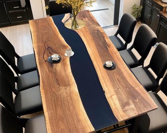 May include: A rectangular dining table featuring two natural live-edge walnut wood slabs joined by a central dark navy blue resin river. The table is surrounded by eight black chairs and holds two small white cups on saucers and a glass vase.