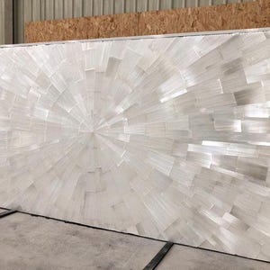 May include: A large rectangular wall panel featuring a sunburst pattern composed of numerous white, translucent selenite crystal tiles. The tiles are arranged in a radiating design that originates from a central point, creating a reflective surface.