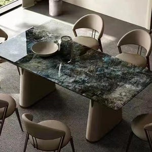 Puede incluir: Una mesa de comedor rectangular con una superficie oscura y veteada y una base de pedestal beige. Seis sillas beige con respaldos curvos y patas de metal oscuro rodean la mesa. Un cuenco y un tarro de cristal están sobre la mesa.