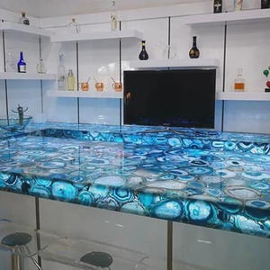 Op de afbeelding: Een moderne keukeneiland met een blauwe agaat aanrechtblad. Het aanrechtblad is verlicht en toont de ingewikkelde patronen van de steen. Witte planken en een zwarte tv zijn aan de muur boven het eiland gemonteerd.