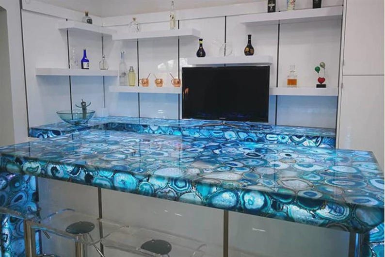 Op de afbeelding: Een moderne keukeneiland met een blauwe agaat aanrechtblad. Het aanrechtblad is verlicht en toont de ingewikkelde patronen van de steen. Witte planken en een zwarte tv zijn aan de muur boven het eiland gemonteerd.