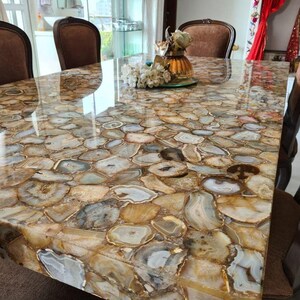 May include: A rectangular dining table with a polished surface made of agate slices in shades of brown, beige, and gray. The table is surrounded by brown upholstered chairs. A vase of flowers sits on the table.