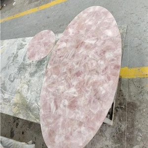 Puede incluir: Una gran encimera de mesa de cuarzo rosa de forma ovalada con una pieza ovalada más pequeña a juego. La superficie tiene un tono rosa claro con un patrón similar a un mosaico. La encimera es probablemente para una mesa de centro o mueble similar.