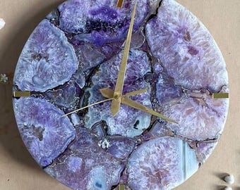 May include: Circular wall clock with a natural amethyst stone face. The clock has gold-coloured hands and hour markers. The purple and white stone has a unique pattern. A decorative piece for any room.