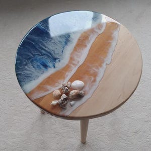 Pode incluir: Mesa lateral redonda de madeira com tampo de resina epóxi com tema de praia. O tampo apresenta um design de oceano azul e branco com efeito de praia arenosa e conchas. A mesa tem três pernas claras.