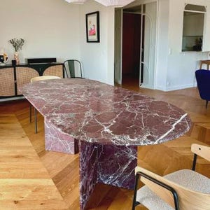 May include: A large, oval-shaped dining table with a burgundy marble top and a matching pedestal base. The table is surrounded by wooden chairs with black metal frames. The room has a hardwood floor and is lit by two pendant lights.
