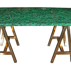 Puede incluir: Mesa rectangular con tapa verde malaquita y patas de caballete color latón. La encimera presenta un patrón arremolinado de verde oscuro y claro. La mesa es un mueble decorativo.