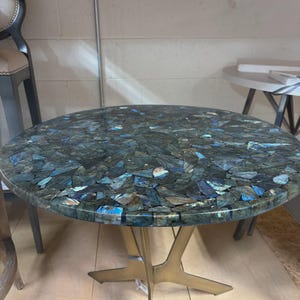 May include: Round coffee table with a mosaic top made of blue, green, and gray stone fragments. The table has a gold-colored metal base with a starburst design. The table is a decorative piece for a living room or lounge area.