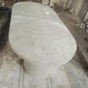 May include: A large, oval-shaped white table with a single, cylindrical pedestal base. The tabletop is constructed from rectangular pieces arranged in concentric circles. The table is a light, neutral color, suitable for various interior design styles.