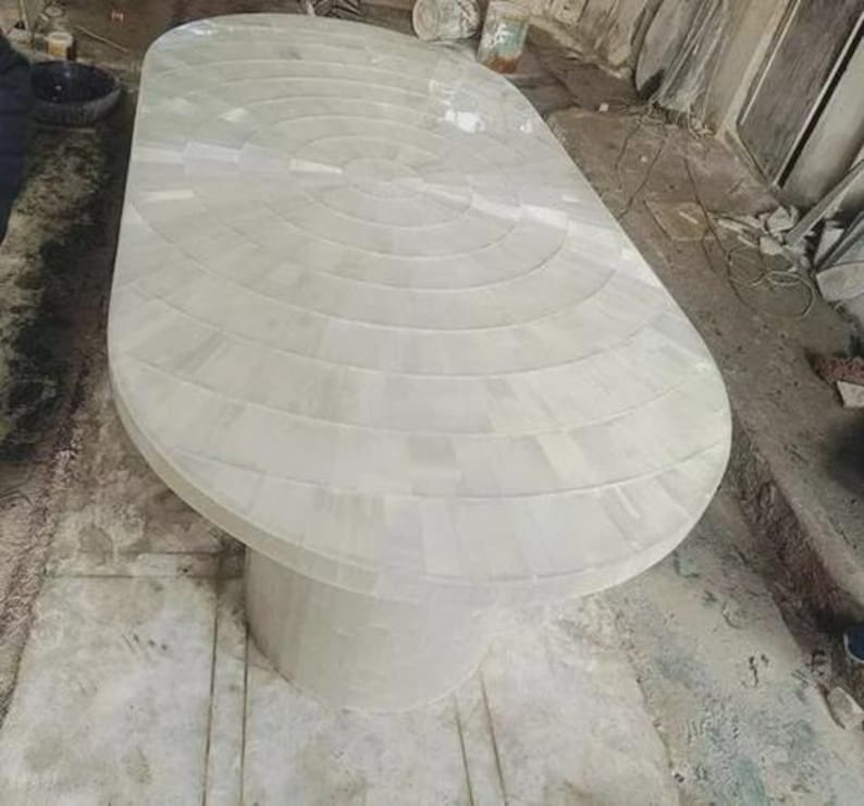 May include: A large, oval-shaped white table with a single, cylindrical pedestal base. The tabletop is constructed from rectangular pieces arranged in concentric circles. The table is a light, neutral color, suitable for various interior design styles.