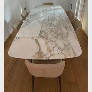 May include: A rectangular marble dining table with a white and gray veined pattern. The table is surrounded by eight beige upholstered chairs. The table and chairs are set on a hardwood floor.