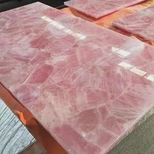 Könnte beinhalten: Eine große, rechteckige Platte aus poliertem Rosenquarz mit einer durchscheinenden, kristallinen Textur in verschiedenen zarten Rosatönen. Die Oberfläche zeigt ein komplexes, gebrochenes Muster aus natürlich verbundenen Steinsegmenten mit glänzendem Finish.