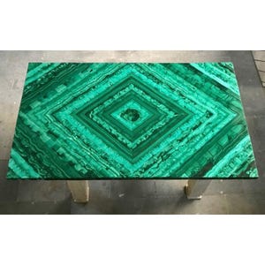 May include: Rectangular malachite table with a striking green diamond pattern. The table is supported by two white pedestals. The malachite has a polished finish, showcasing the natural veining and color variations of the stone.