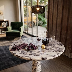 May include: A circular marble table with a white and grey pattern, set with wine glasses, a decanter, grapes, and a candle. A dark green velvet armchair and a hanging light fixture are in the background.