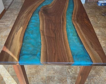 Può includere: Tavolo rettangolare in legno con un fiume di resina turchese al centro. Il tavolo ha venature del legno marrone scuro e quattro robuste gambe in legno marrone. La resina sembra essere larga circa 76 cm.