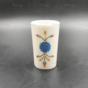 May include: A white marble tumbler with a floral design. The design features a blue flower with orange, blue, and green accents. The tumbler is cylindrical and stands upright on a dark surface.