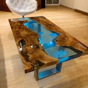 Puede incluir: Una mesa de centro rectangular con un diseño único. La mesa presenta una tapa de madera con borde natural, que rodea una sección central llena de resina azul vibrante. La mesa está soportada por una base metálica.